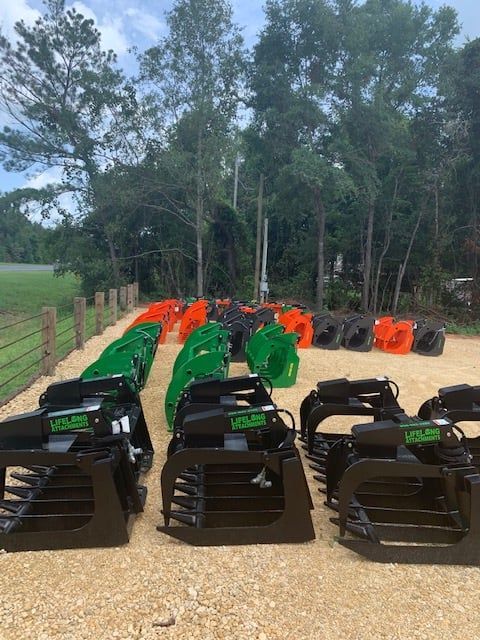 Rows of heavy-duty tractor grapple attachments in various colors on gravel, backed by trees and a fence.