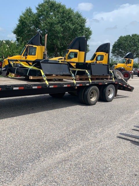Four yellow truck cabs on a flatbed trailer, secured with straps. Asphalt ground, trees in background.