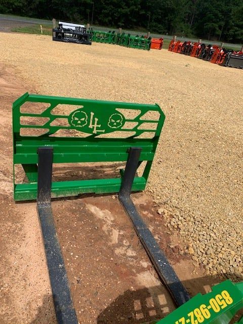 Green forklift attachment with forks, logo, on a gravel lot with tractors.