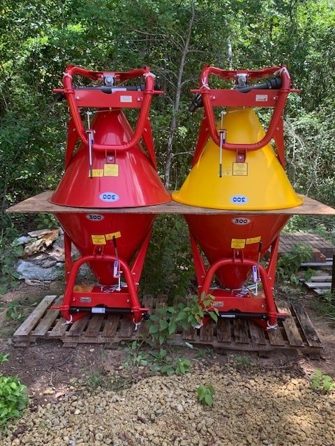 Two agricultural fertilizer spreaders, red and yellow, on pallets in a wooded outdoor area.