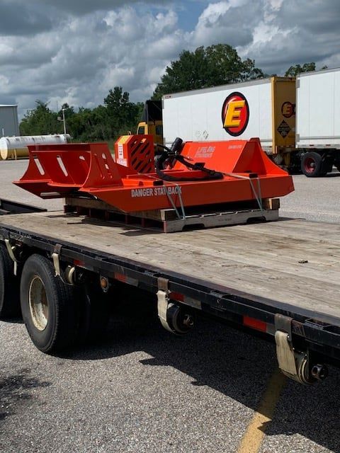 Orange brush cutter on a flatbed trailer, in front of white trailers and a cloudy sky.