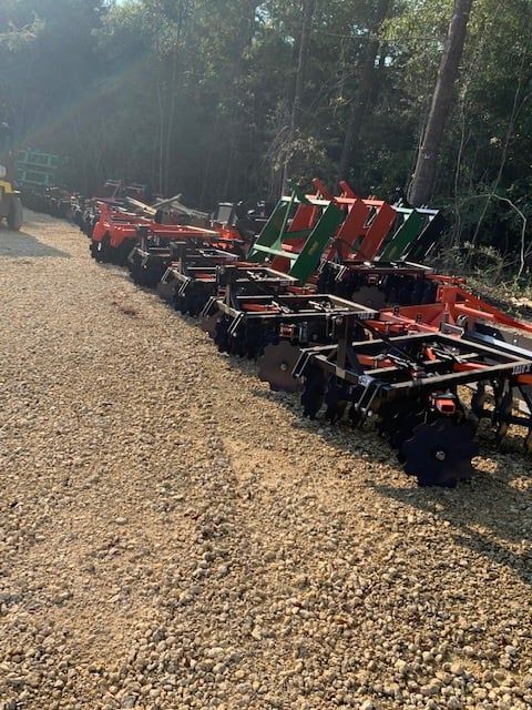 Farm equipment lined up outdoors on gravel. Various colors: orange, green, black. Sunny setting.