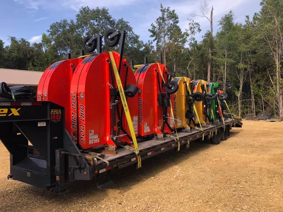 Tractor attachments on a flatbed trailer, secured with straps. Various colors: red, yellow, and green. Outdoors.