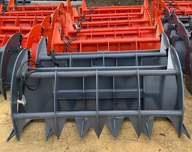 Gray and orange grapple buckets for heavy machinery are arranged outside.