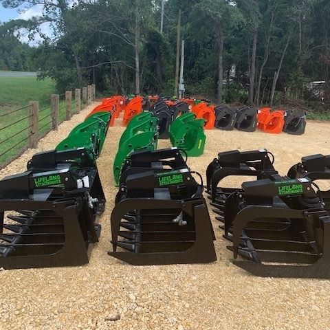 Rows of new, black, green, orange, and gray grapple buckets on gravel, outdoors.