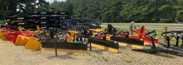 Farm implements, including box blades and tillers, are displayed outside on gravel. Green trees in the background.
