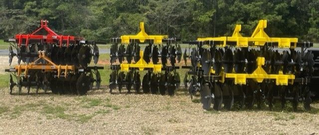 Farm equipment, including disc harrows, of various sizes, in a grassy area with trees in the background.
