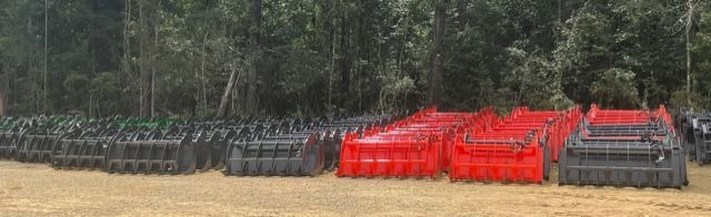Rows of dumpsters, including red and dark grey containers, sit in a field with a treeline in the background.