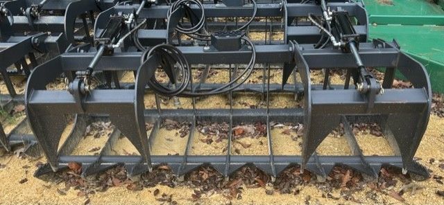 A large, dark metal grapple attachment with tines and a curved top, resting on soil and debris.