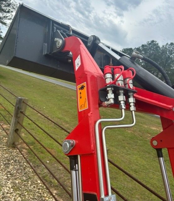 Red tractor front loader with hydraulic lines and a black bucket. Outdoors, grassy slope in the background.
