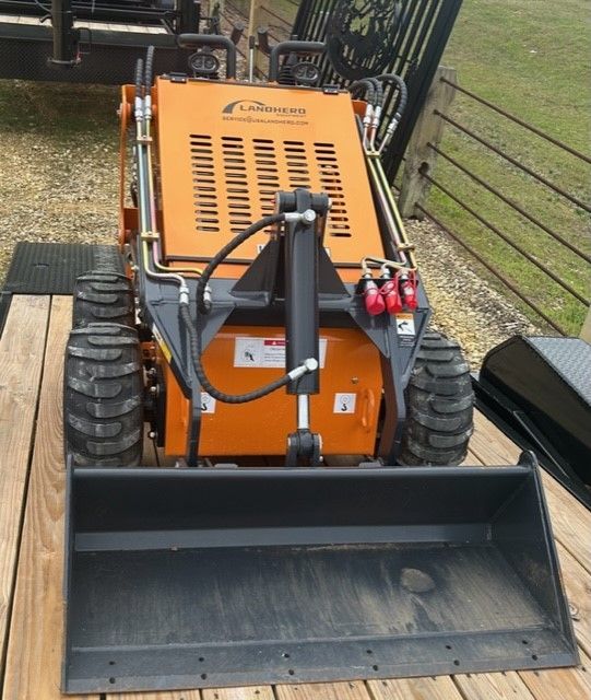 Orange Landscaper Pro mini skid steer with a black bucket on a wooden trailer.