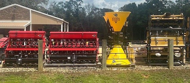 Agricultural machinery, including red and yellow planters, are lined up in front of a building and trees.