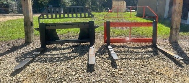 Two forklift attachments, one black, one red, resting on gravel.