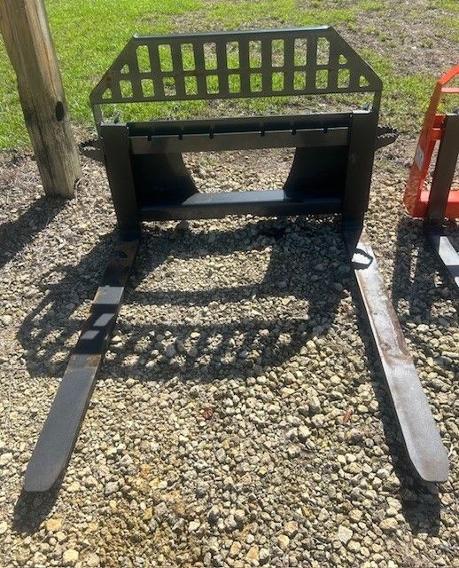 Black pallet fork attachment on gravel; green grass background.