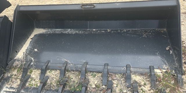 A black front-end loader bucket resting on the ground with grass and dirt.