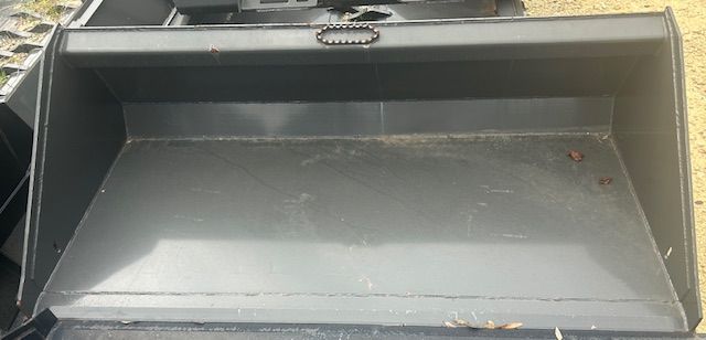 An empty, dark gray metal bucket, possibly for a loader or tractor, sits on the ground.