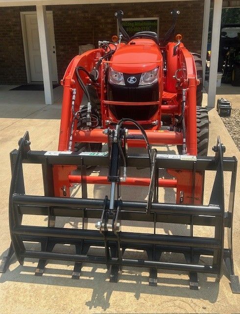 Orange Kubota tractor with grapple attachment, parked outside on concrete.