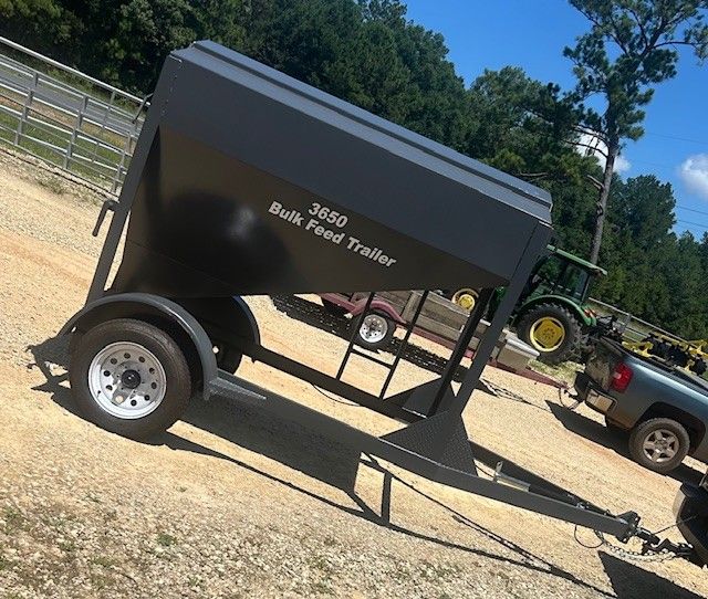 A black 3650 bulk feed trailer on a gravel surface with a farm in the background.