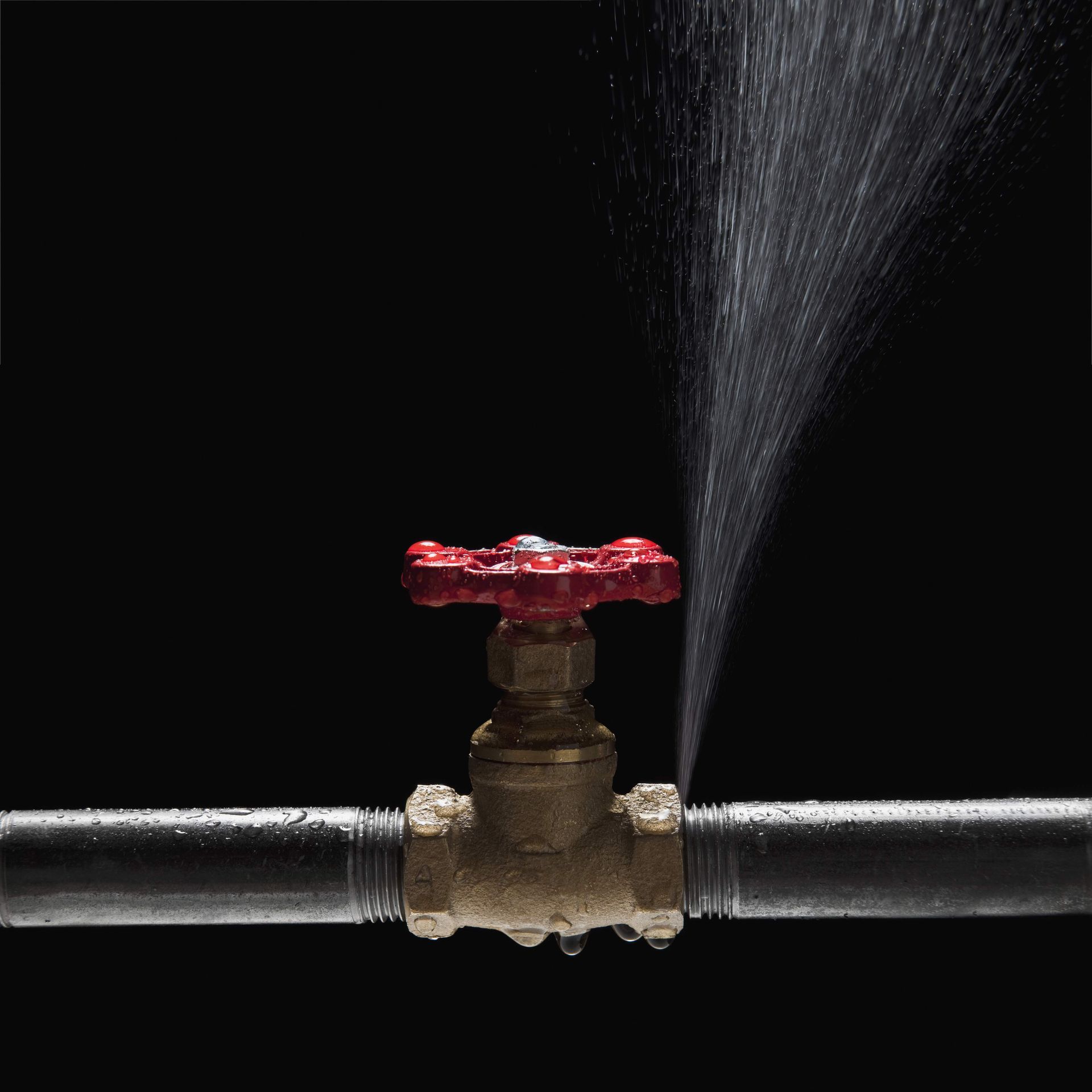 Water is spraying from a leaking pipe valve with a red handle.