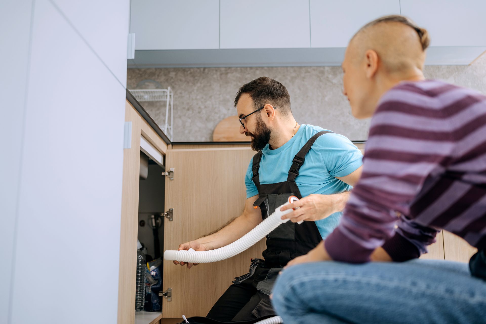Plumber working under the kitchen sink while a client looks on. Plumber working under the kitchen sink while a client looks on.