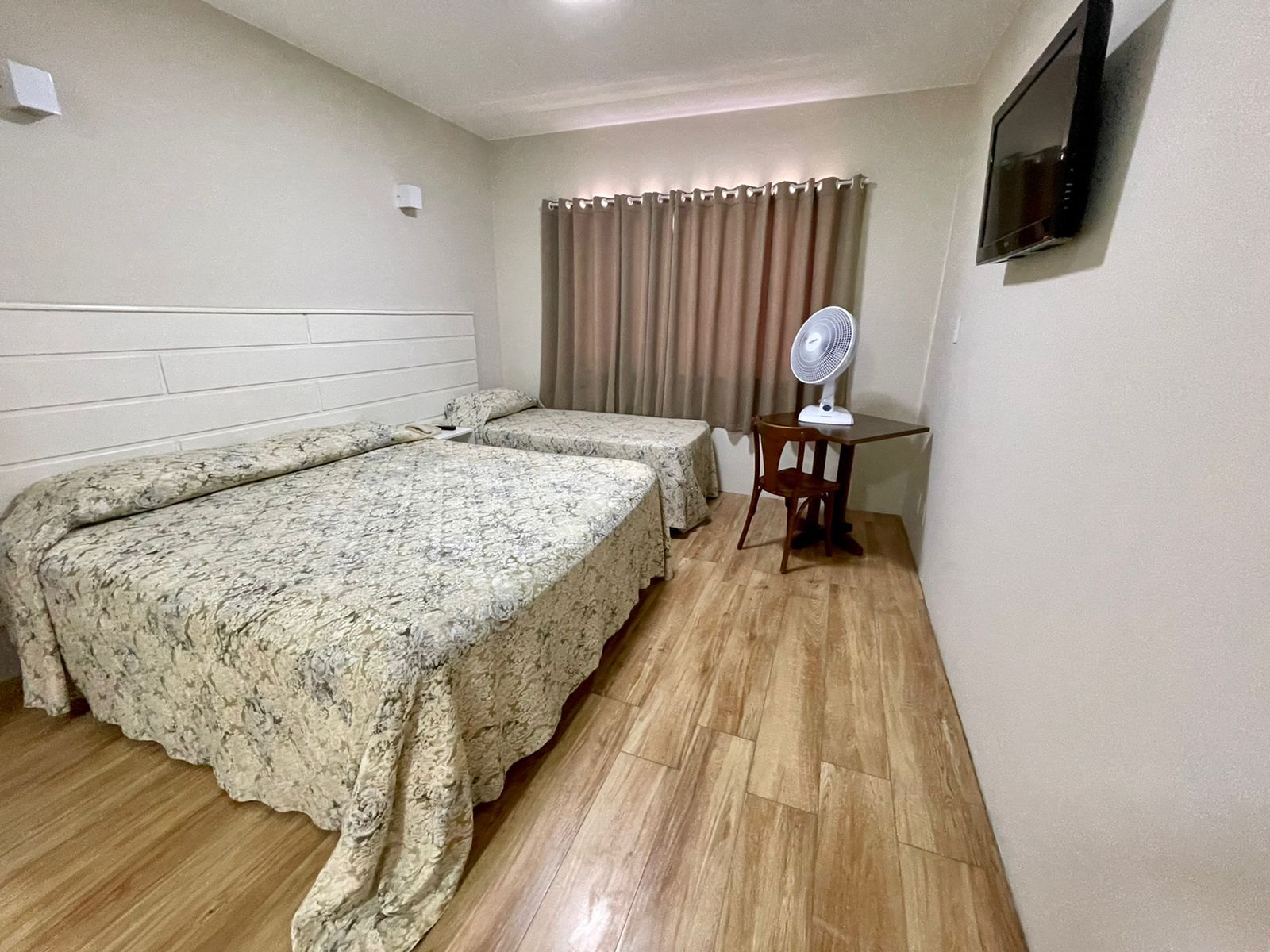 Um quarto de hotel com duas camas, uma pequena mesa com um ventilador e uma TV montada.