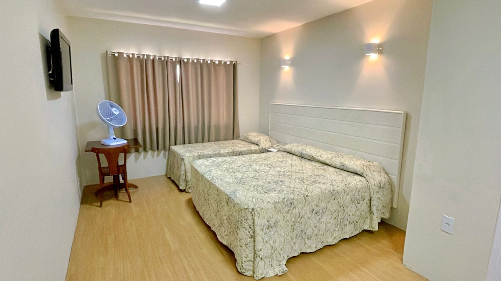 Quarto com duas camas de solteiro, cortinas bege, mesa pequena com ventilador e TV na parede.