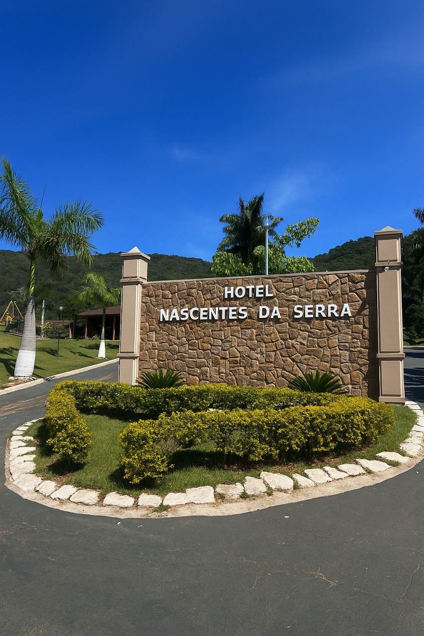 Placa do Hotel Nascentes da Serra, entrada de pedra, arbustos verdes, céu azul.