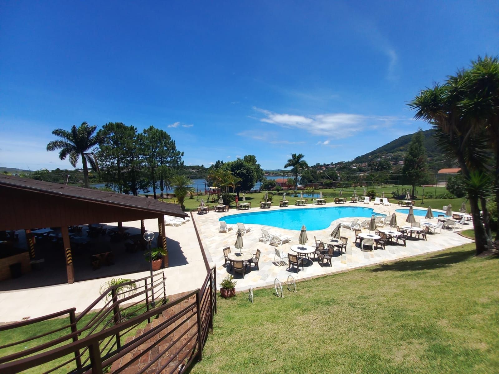 Área de piscina com mesas e guarda-sóis, com vista para um lago e colinas verdes sob um céu azul.