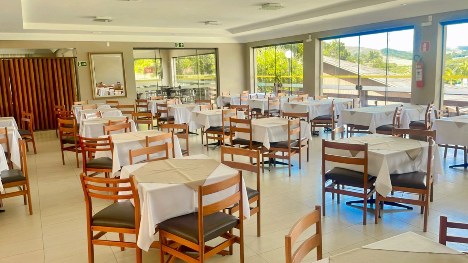 Restaurante vazio com mesas postas para refeições, de frente para grandes janelas com vista para uma paisagem.