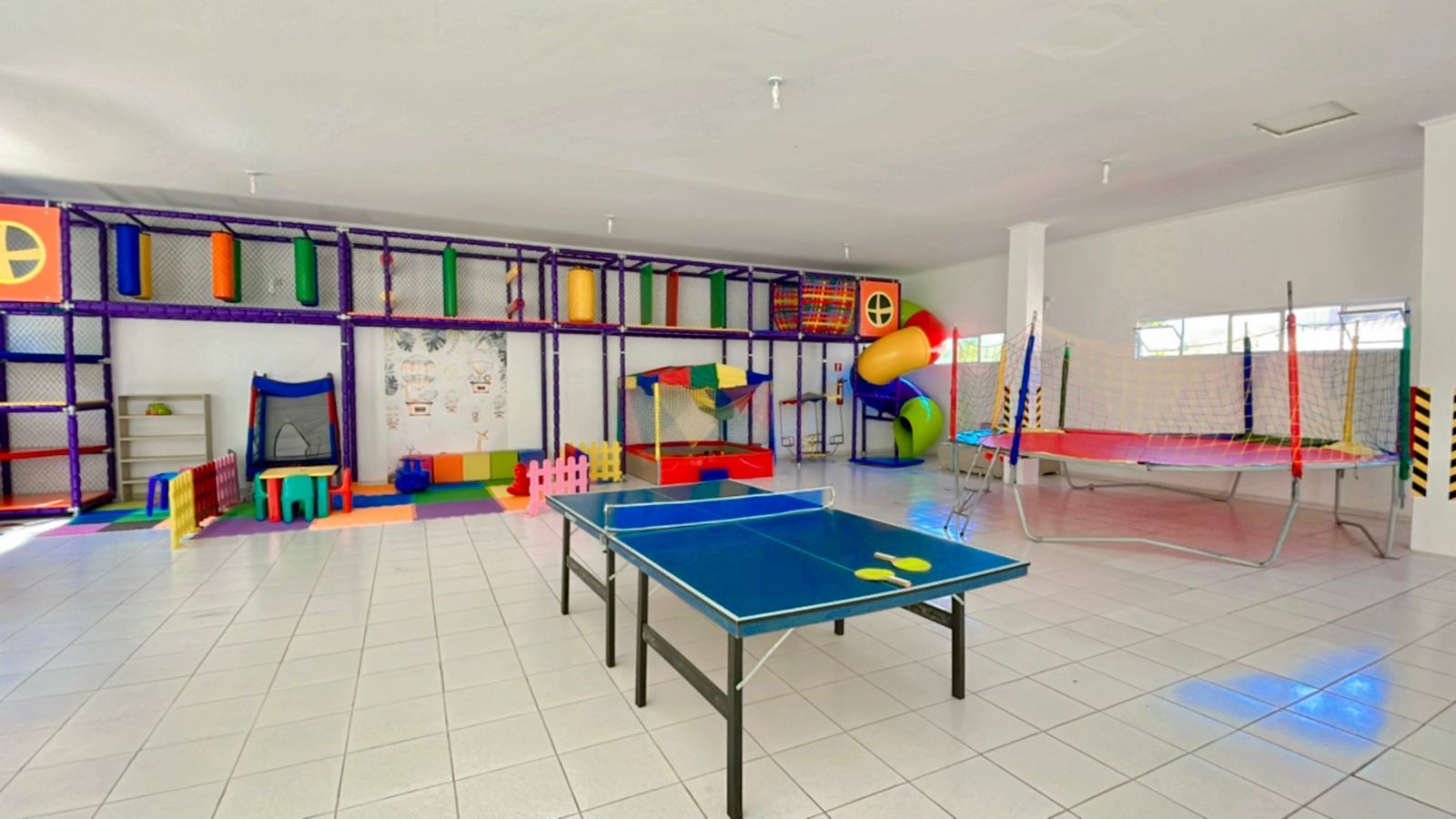 Sala de jogos interna com mesa de pingue-pongue, estruturas de escalada e trampolim.
