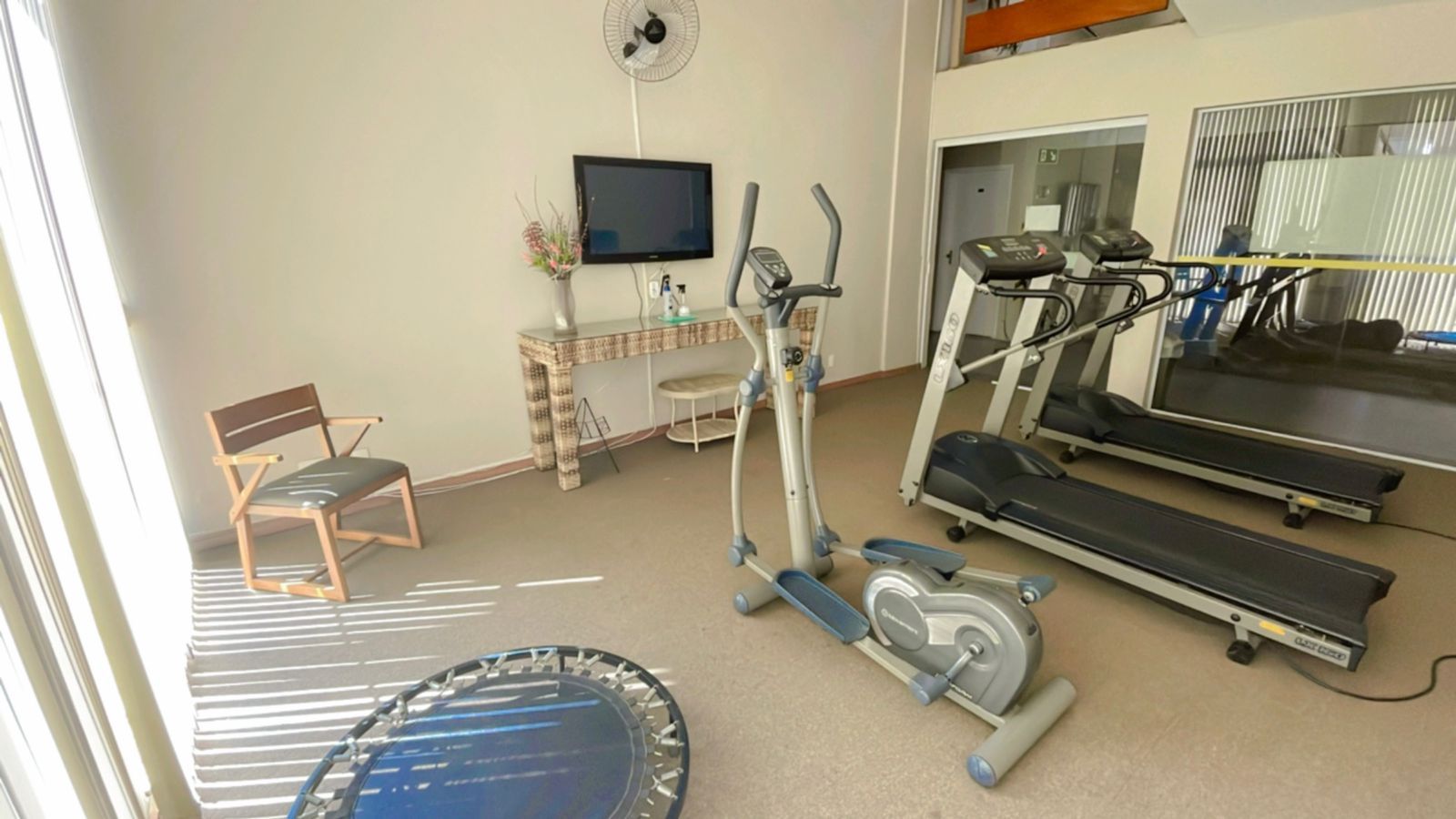 Academia com aparelhos de ginástica, cadeira, televisão e ventilador.