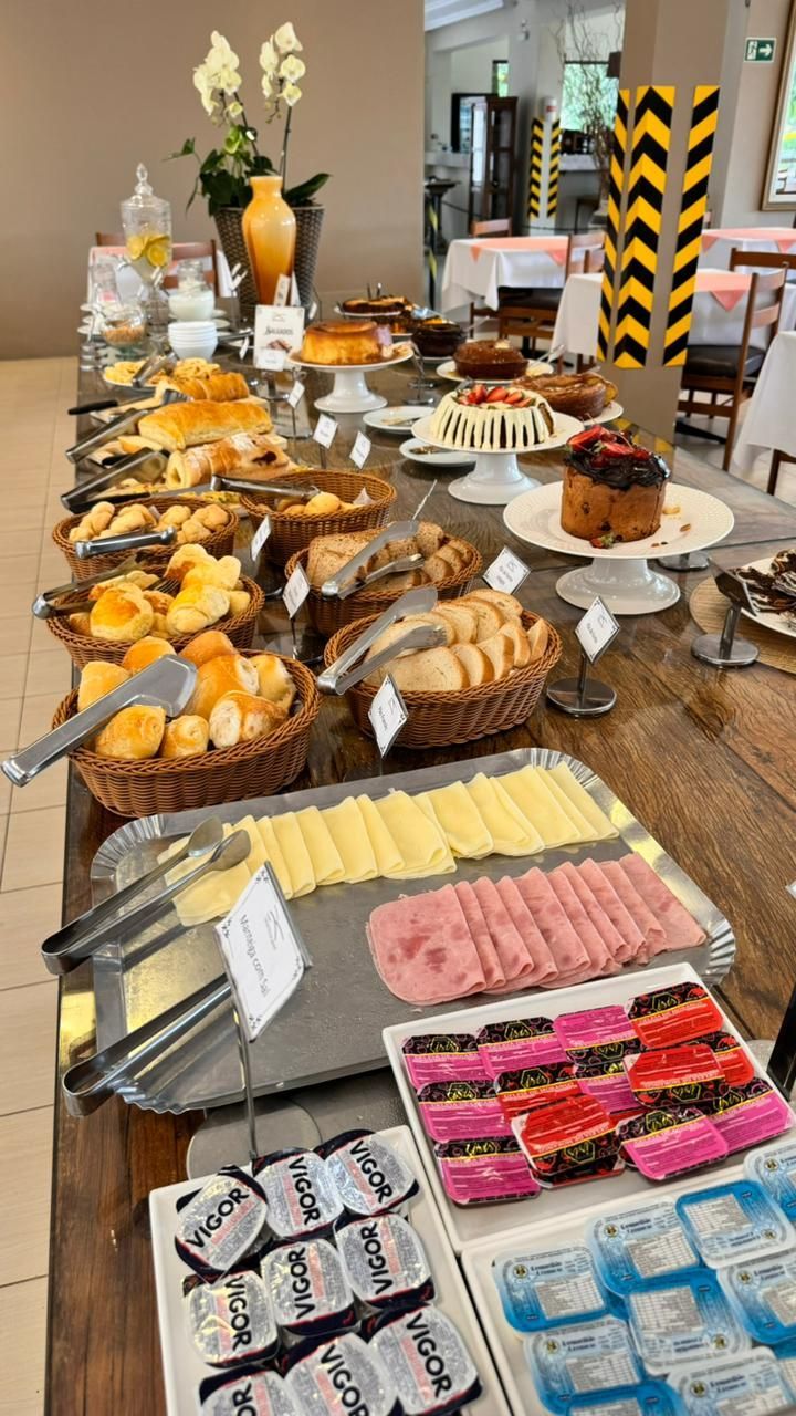 Buffet de café da manhã com doces, queijos, frios e condimentos em uma longa mesa de madeira.