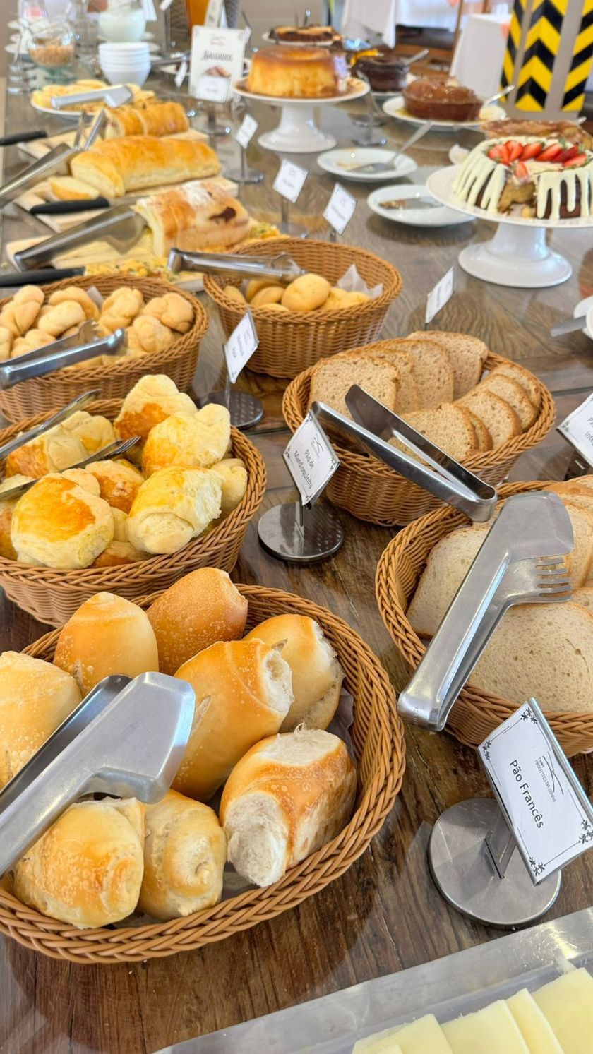Buffet de café da manhã com diversos pães, queijos, carnes e doces em uma mesa de madeira.
