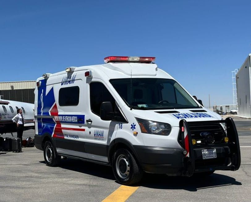 Ambulancia blanca y azul en la pista de un aeropuerto con un jet privado y una persona al fondo bajo un cielo azul.