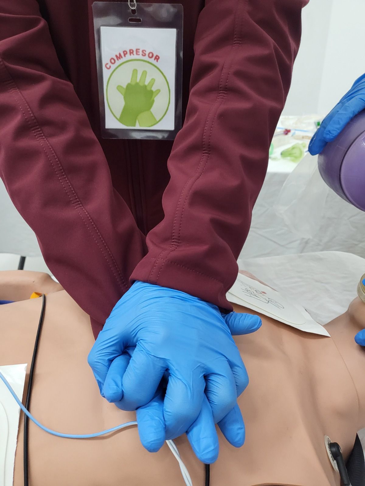 Persona con guantes azules realizando compresiones torácicas en un maniquí. El cartel dice 
