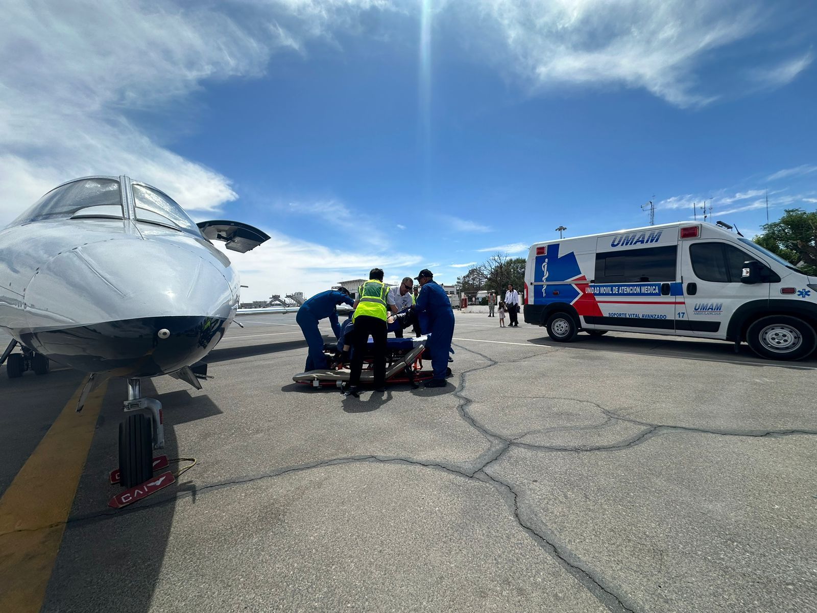 Personal médico asiste a alguien cerca de una avioneta y una ambulancia en una pista bajo un cielo azul.