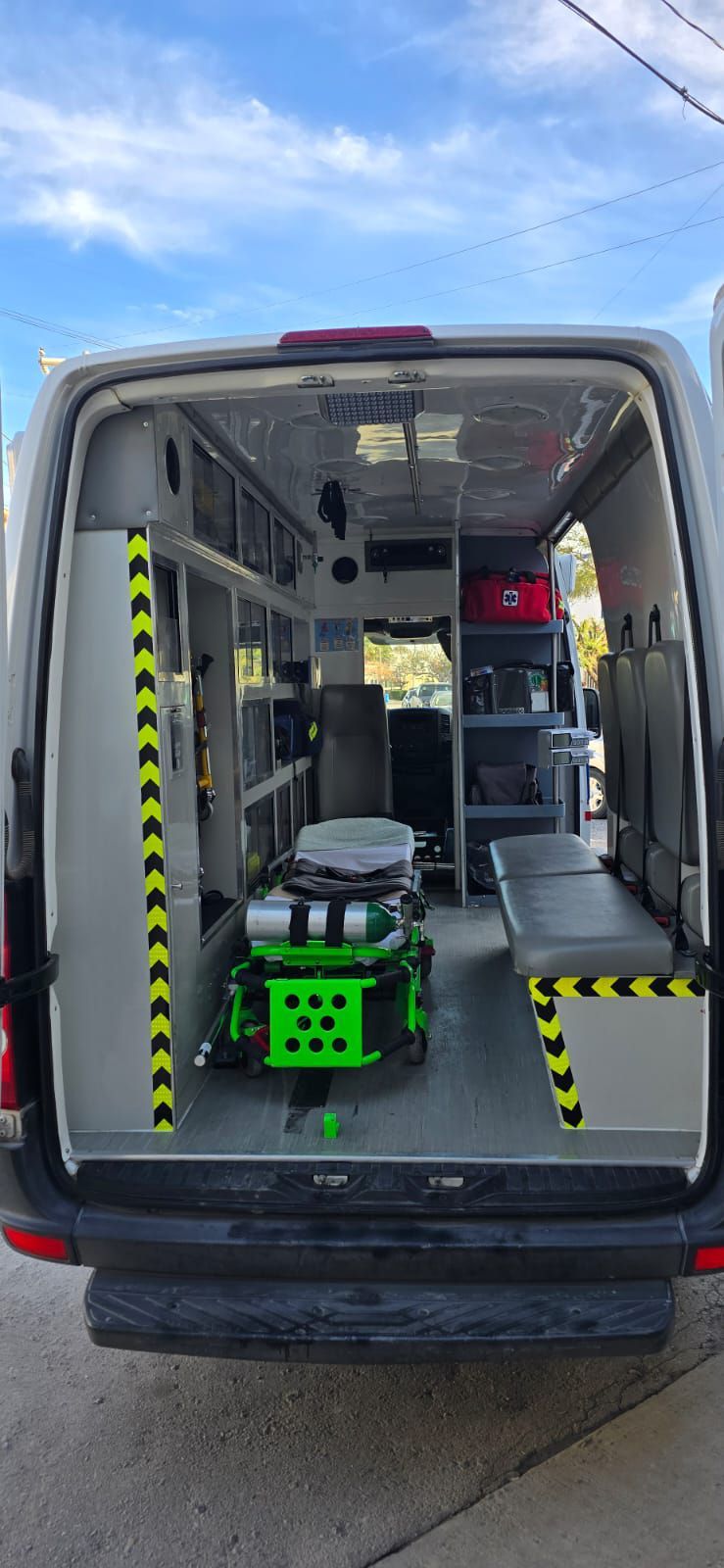 Interior de una ambulancia. Una camilla verde se encuentra dentro de la furgoneta gris. Se ven estantes y un banco.