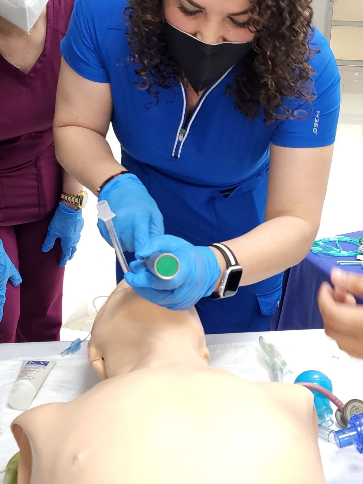 Profesional médica que muestra la intubación en un maniquí. Lleva uniforme azul, mascarilla y guantes.