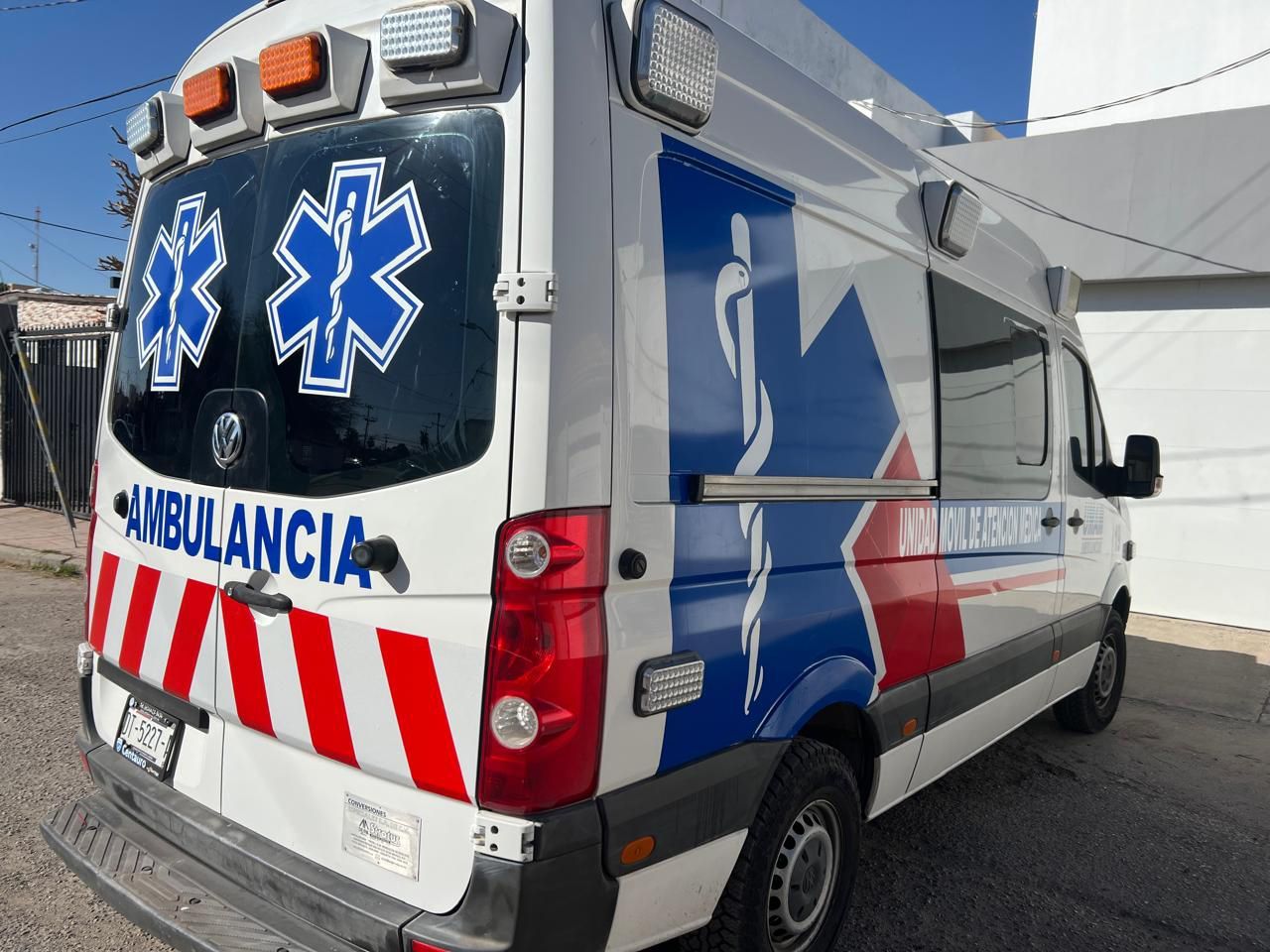 Ambulancia blanca con símbolos médicos azules y franjas rojas y blancas. Texto: 