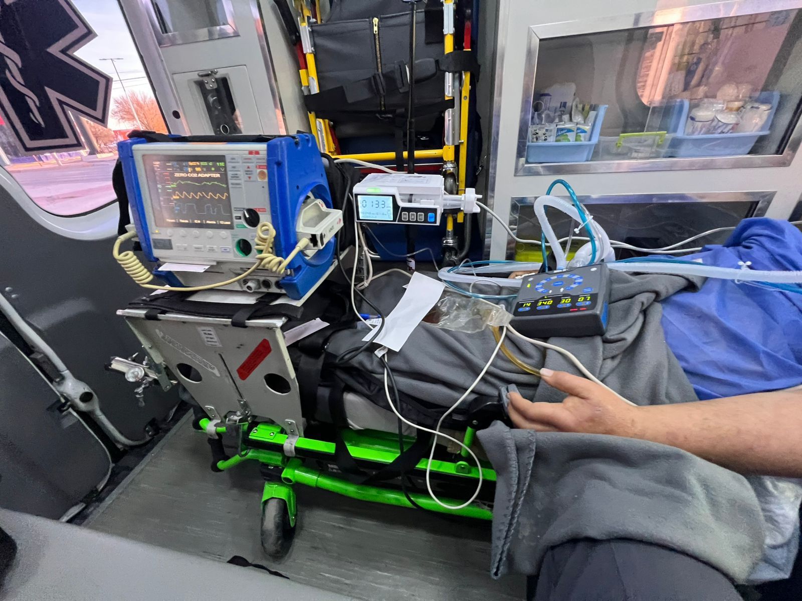 Dentro de una ambulancia, el equipo médico rodea a un paciente en una camilla. Una mano descansa sobre una manta gris.