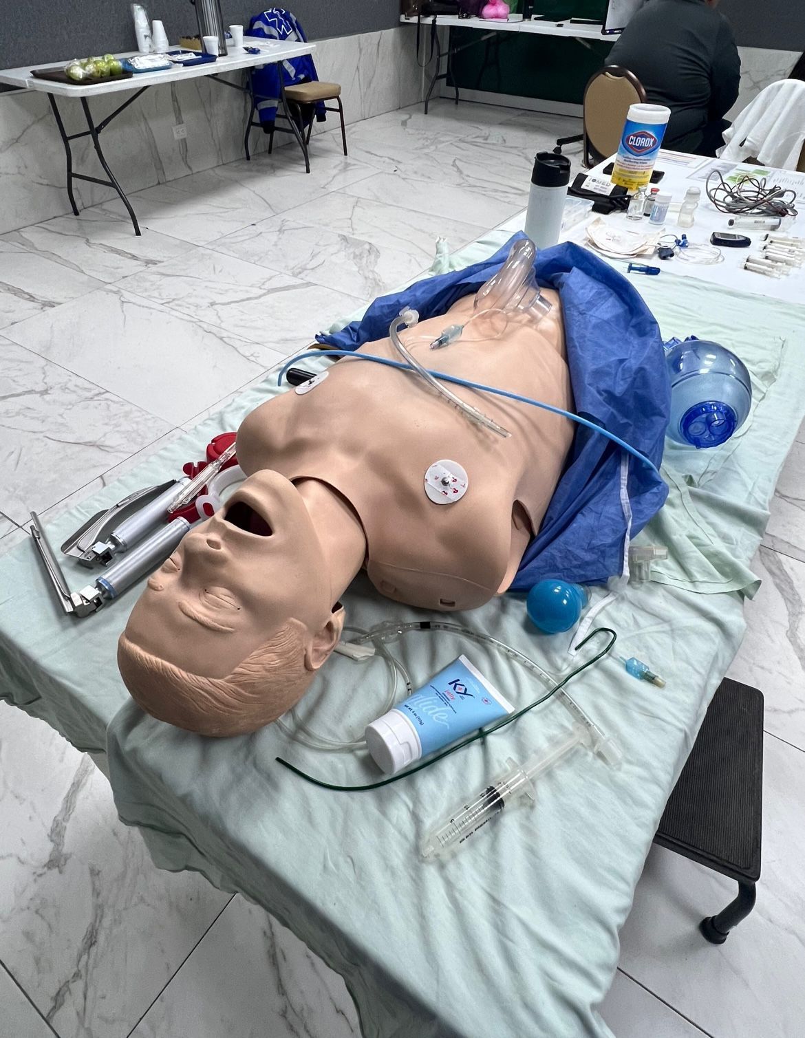 Maniquí de prácticas médicas sobre una mesa con material médico. En una sala con mesas y una persona.