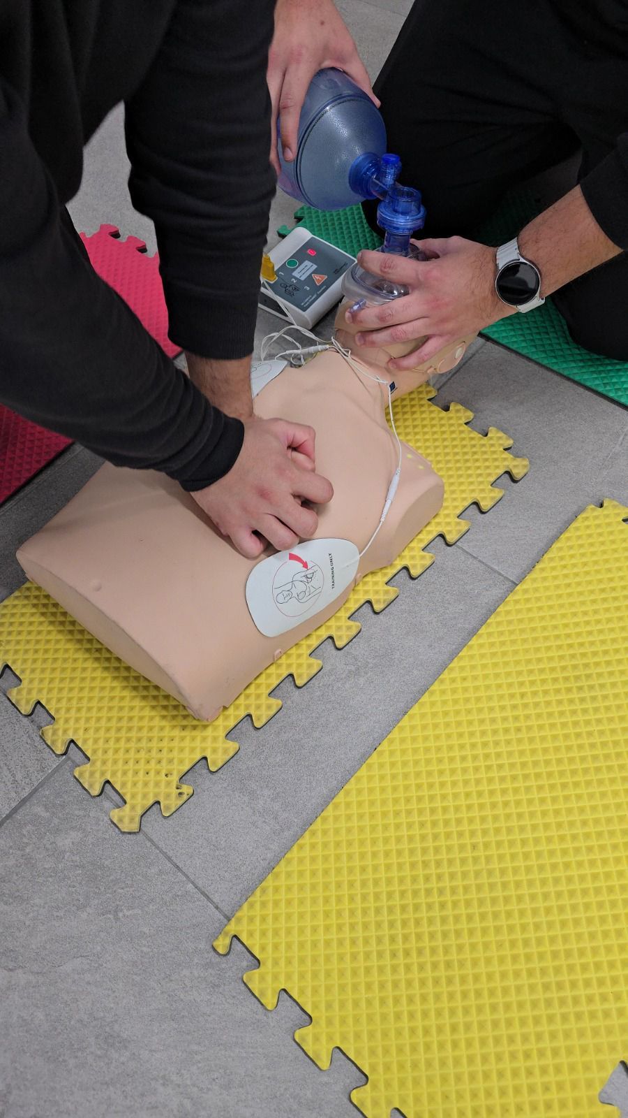 Dos personas realizando RCP en un maniquí, una con compresiones torácicas y la otra con una bolsa-máscara con válvula.