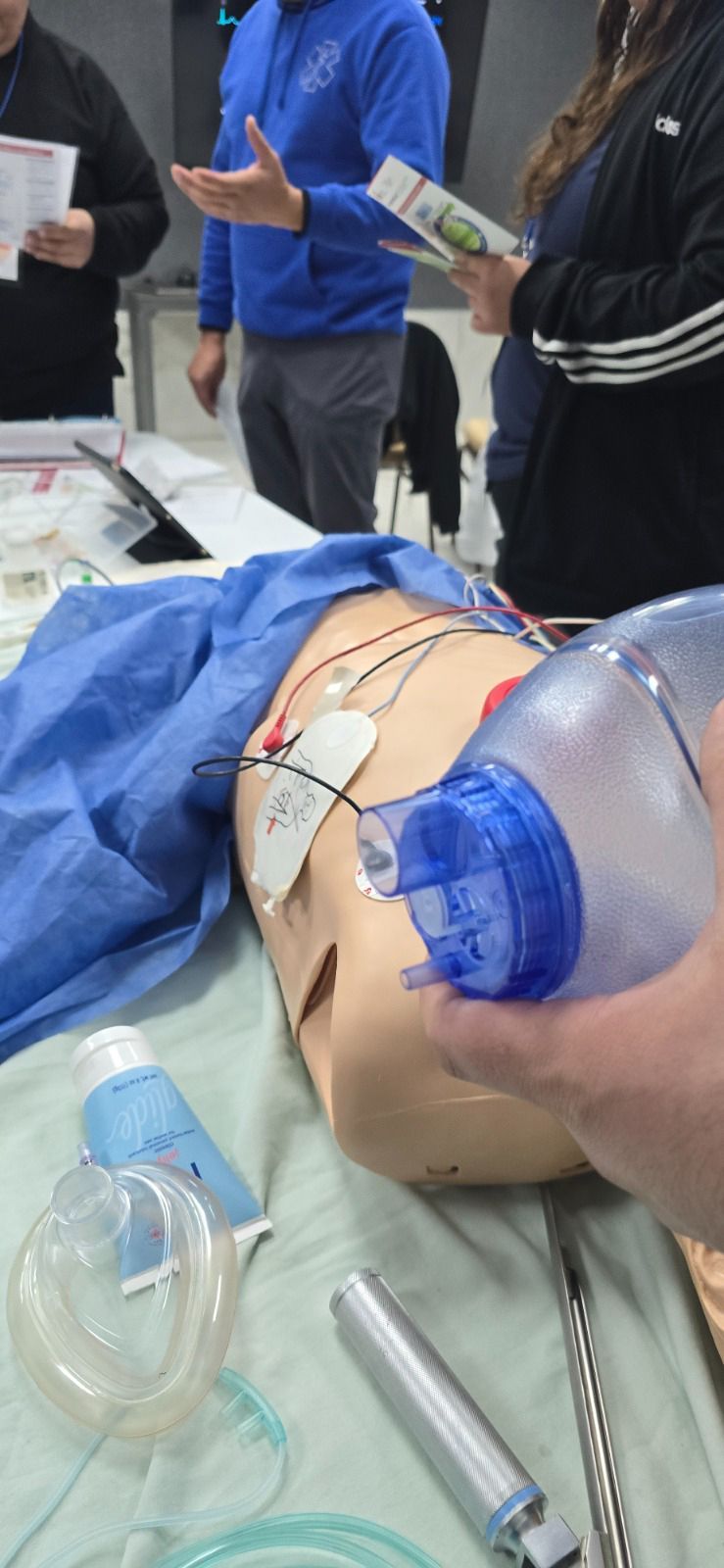 Entorno de formación médica con un maniquí en uso. Una persona sostiene una mascarilla y otros equipos visibles.