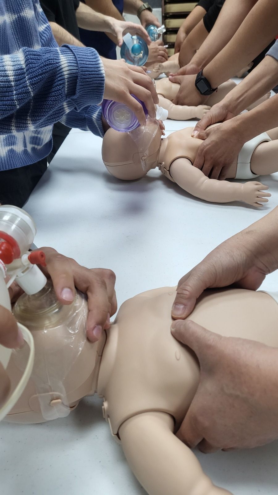 Manos demostrando técnicas de RCP en maniquíes infantiles. Varios participantes participan en el ejercicio en una mesa.