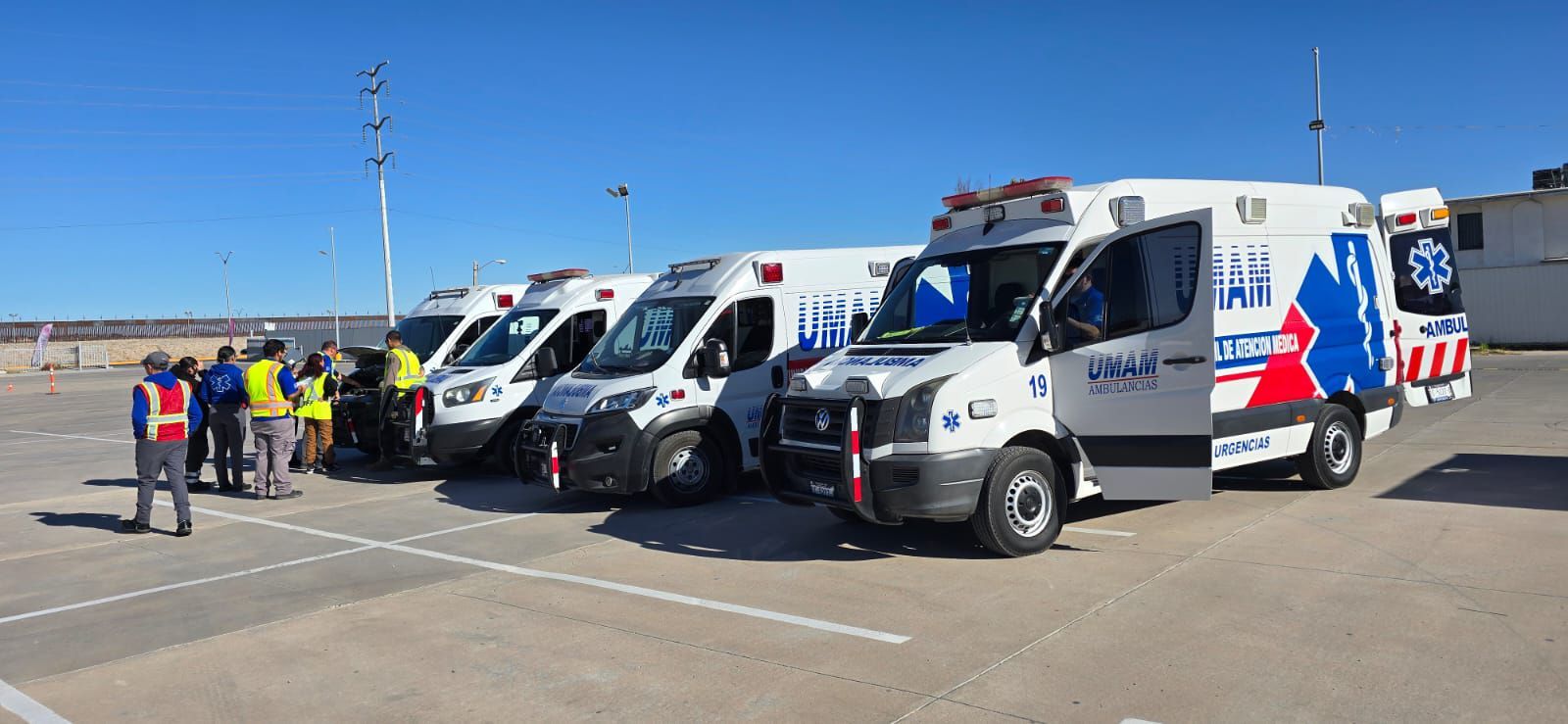 Una fila de ambulancias blancas con las puertas abiertas, estacionadas sobre asfalto. Hay gente cerca.