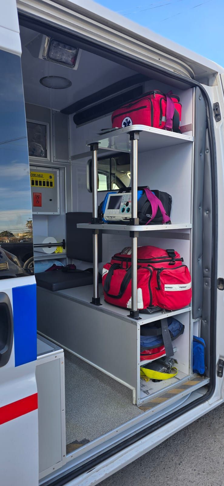 Dentro de una ambulancia con suministros médicos. Se ven bolsas rojas, equipo médico y estantes.