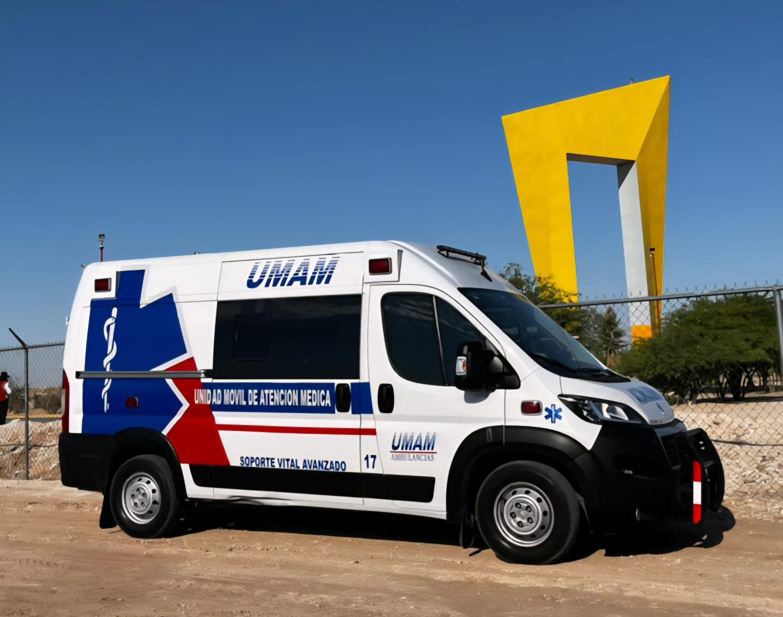 Ambulancia blanca con detalles en azul y rojo, estacionada al aire libre cerca de una escultura amarilla. Se ve la inscripción 