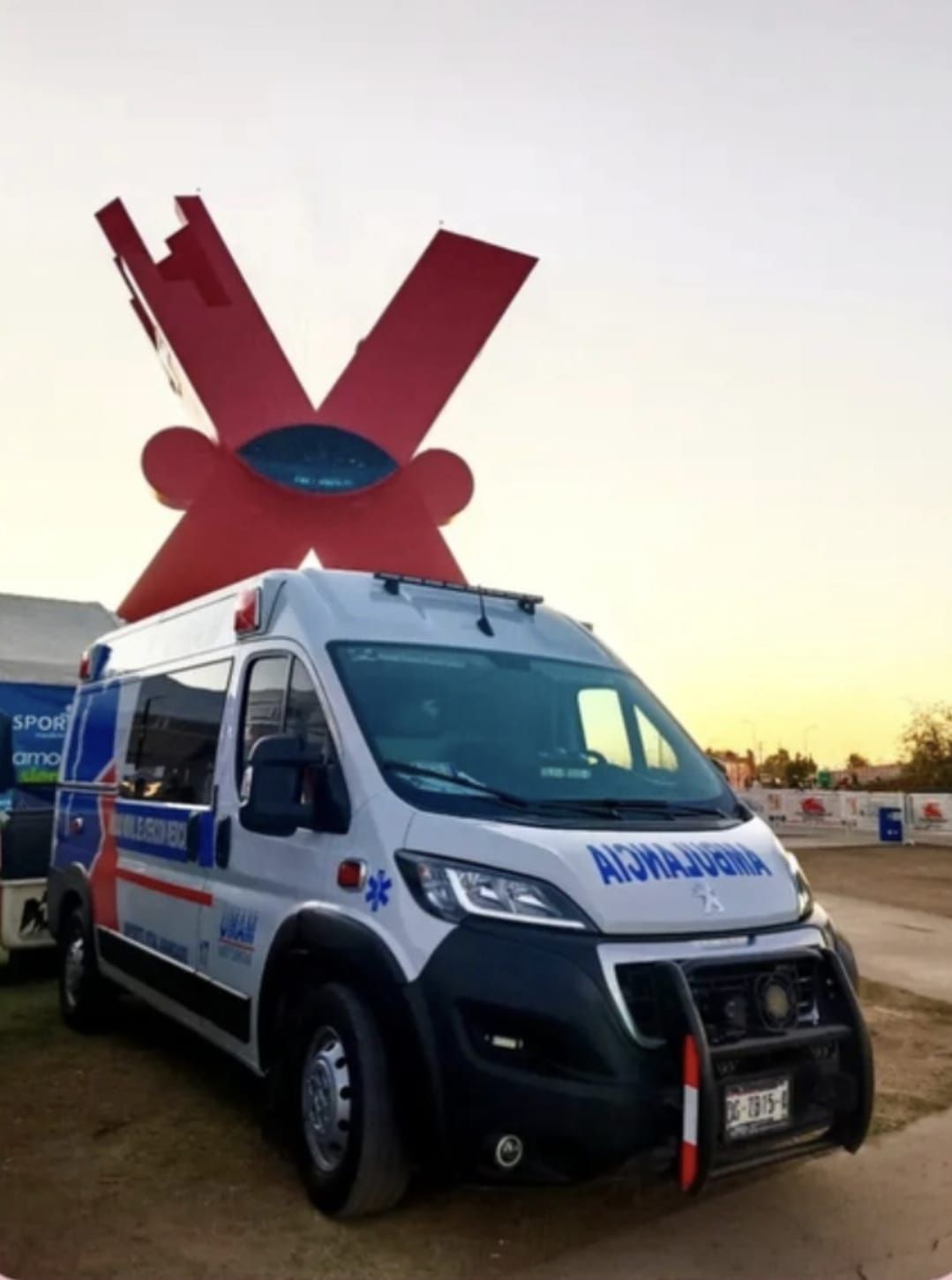 Ambulancia estacionada frente a una gran escultura de una X roja; vehículo azul y blanco, entorno al aire libre.