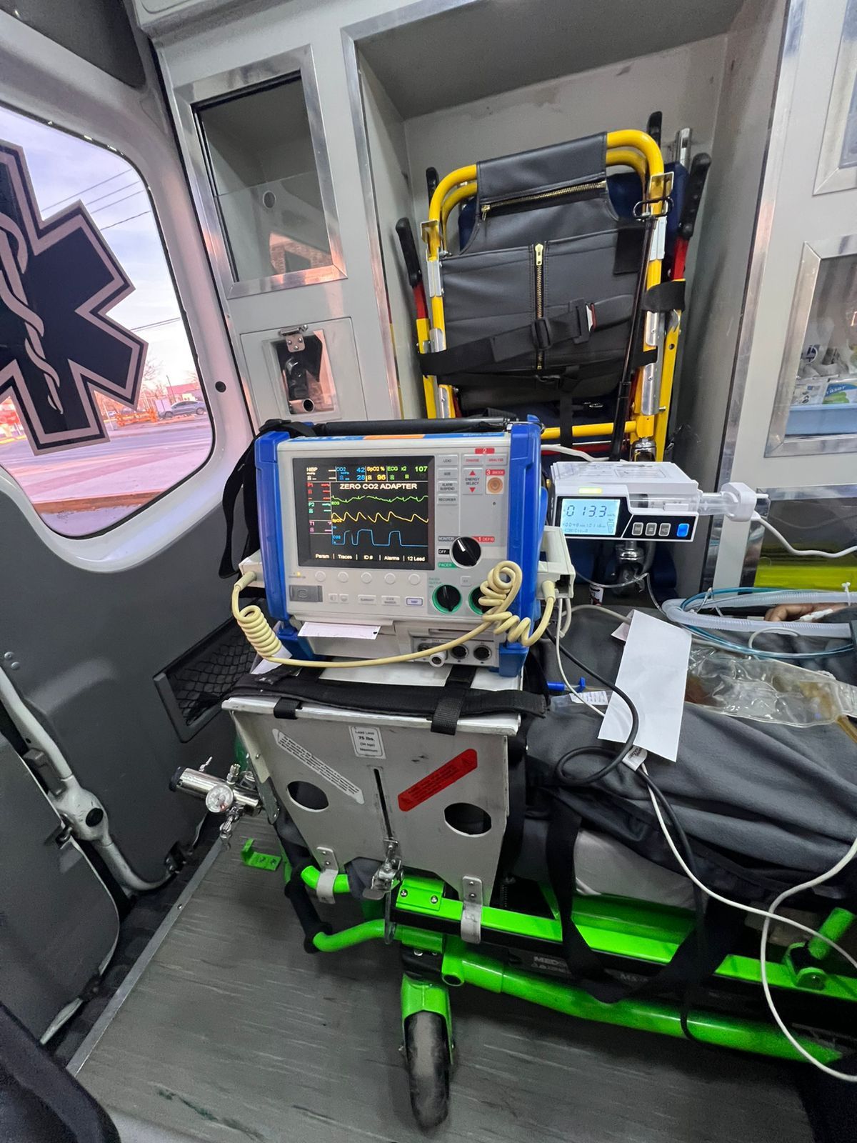 Dentro de la ambulancia, monitor/desfibrilador sobre soporte metálico; camilla al fondo.