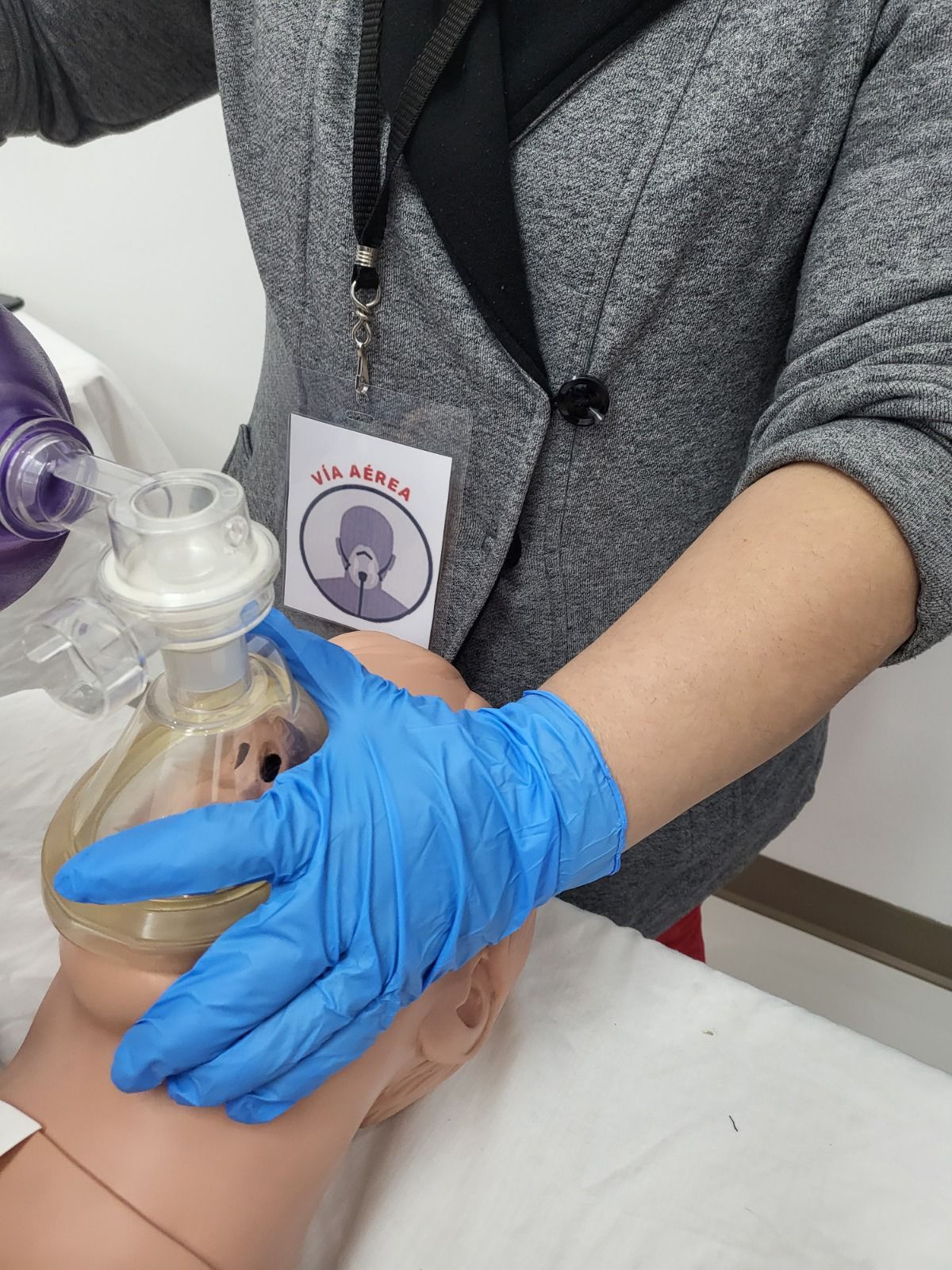 Persona con chaqueta gris muestra el uso de una mascarilla con válvula de bolsa en un maniquí. Una mano con guante azul sostiene la mascarilla.